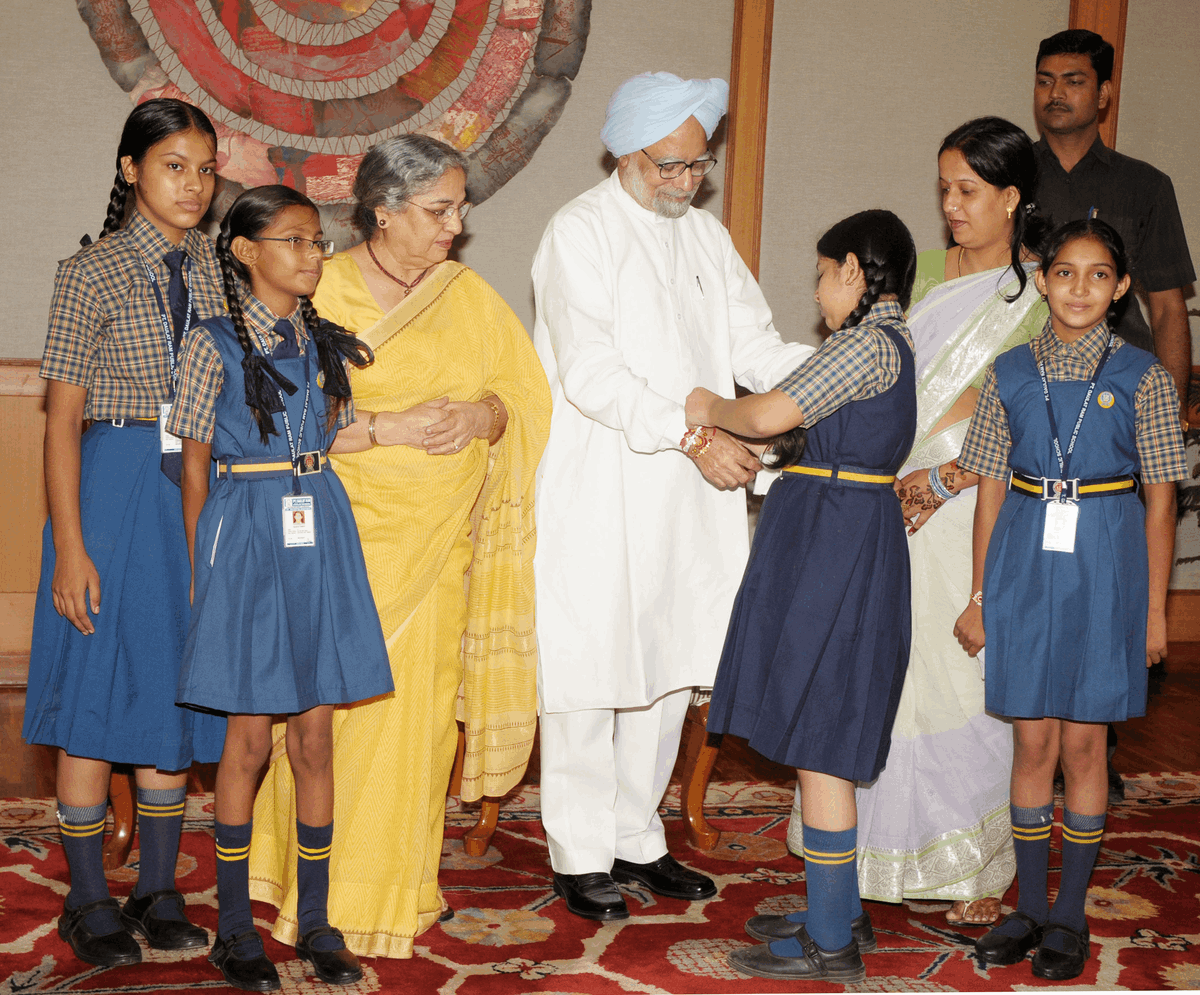 criancas-amarrando-rakhi-primeiro-ministro-manmohan-singh-2011 Crianças em idade escolar amarram rakhi no pulso do primeiro-ministro da Índia, Dr. Manmohan Singh, durante o Raksha Bandhan em Nova Délhi, em 2011, com Gursharan Kaur presente